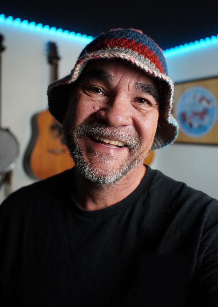 Michael "Mash" Ashley, smiling in a crocheted bucket hat, guitars on the wall behind him